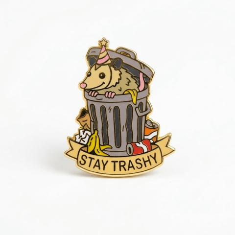 An enamel pin featuring an opossum wearing a pink striped party hat, popping out of a grey trash can surrounded by banana peels and crushed soda cans. A banner across the bottom reads "STAY TRASHY".