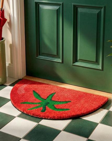 A half-circle red tomato doormat with a green stem design on a green and white checkered tile floor in front of a green door.