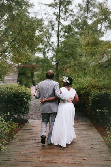 My husband and I walking hand-in-hand with our backs to the camera, down a wooden path surrounded by greenery. Joey is wearing a gray suit and has an arm around my waist. I’m in a simple sleeves, long white dress, with a floral headdress. He is gesturing with his other hand and you can see from my profile that I’m laughing.