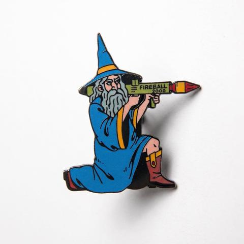 Enamel pin of a long-bearded wizard in a blue robe and pointed hat, kneeling while aiming a rocket launcher labeled "FIREBALL 2000".
